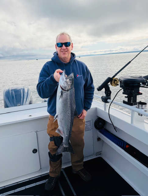 A man holding a large fish he caught on the Kwigwis Adventures boat. Calm waters and cloudy skies are visible in the background.